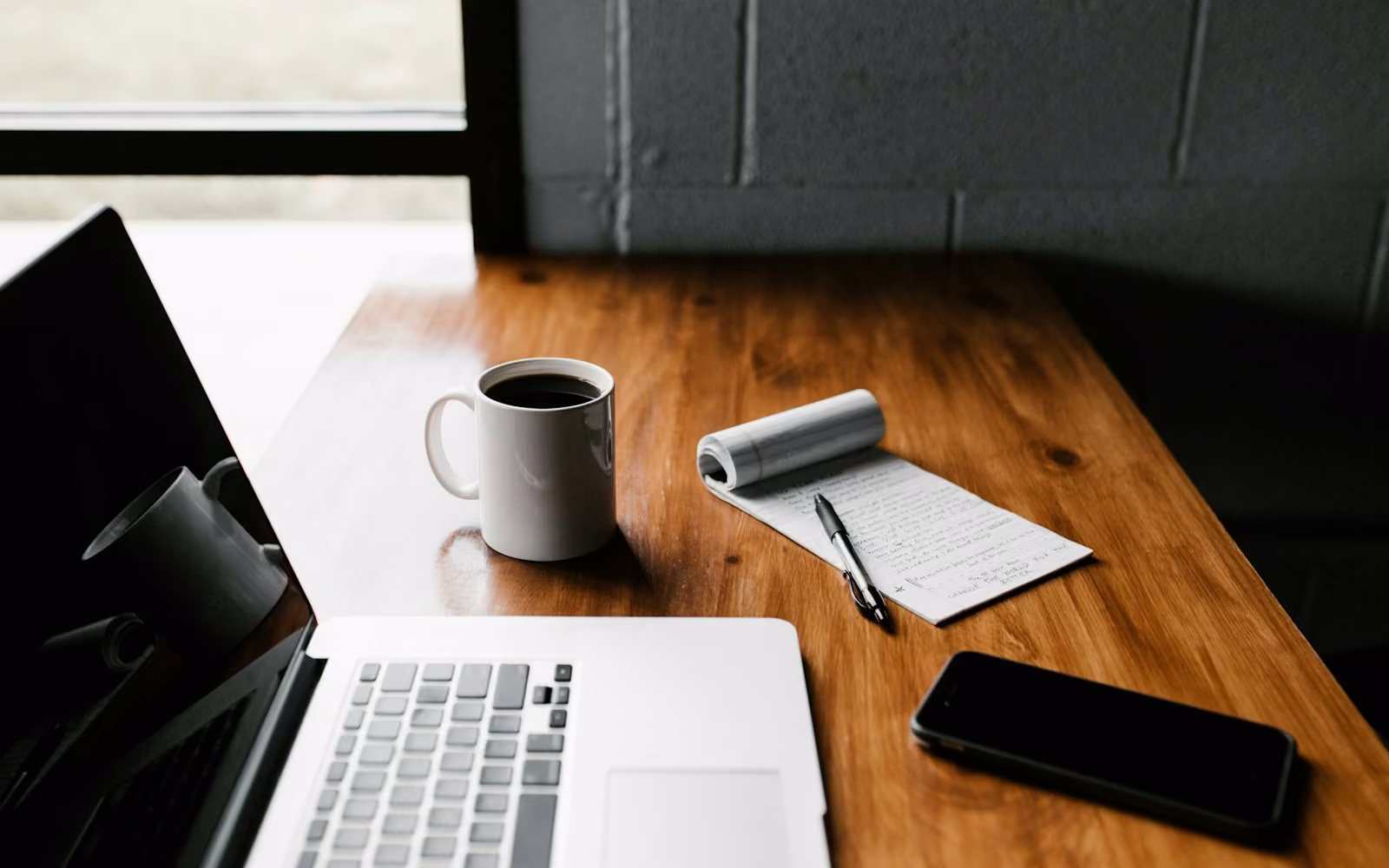 Laptop, buku catatan kecil, pulpen, dan secangkir kopi di meja kayu