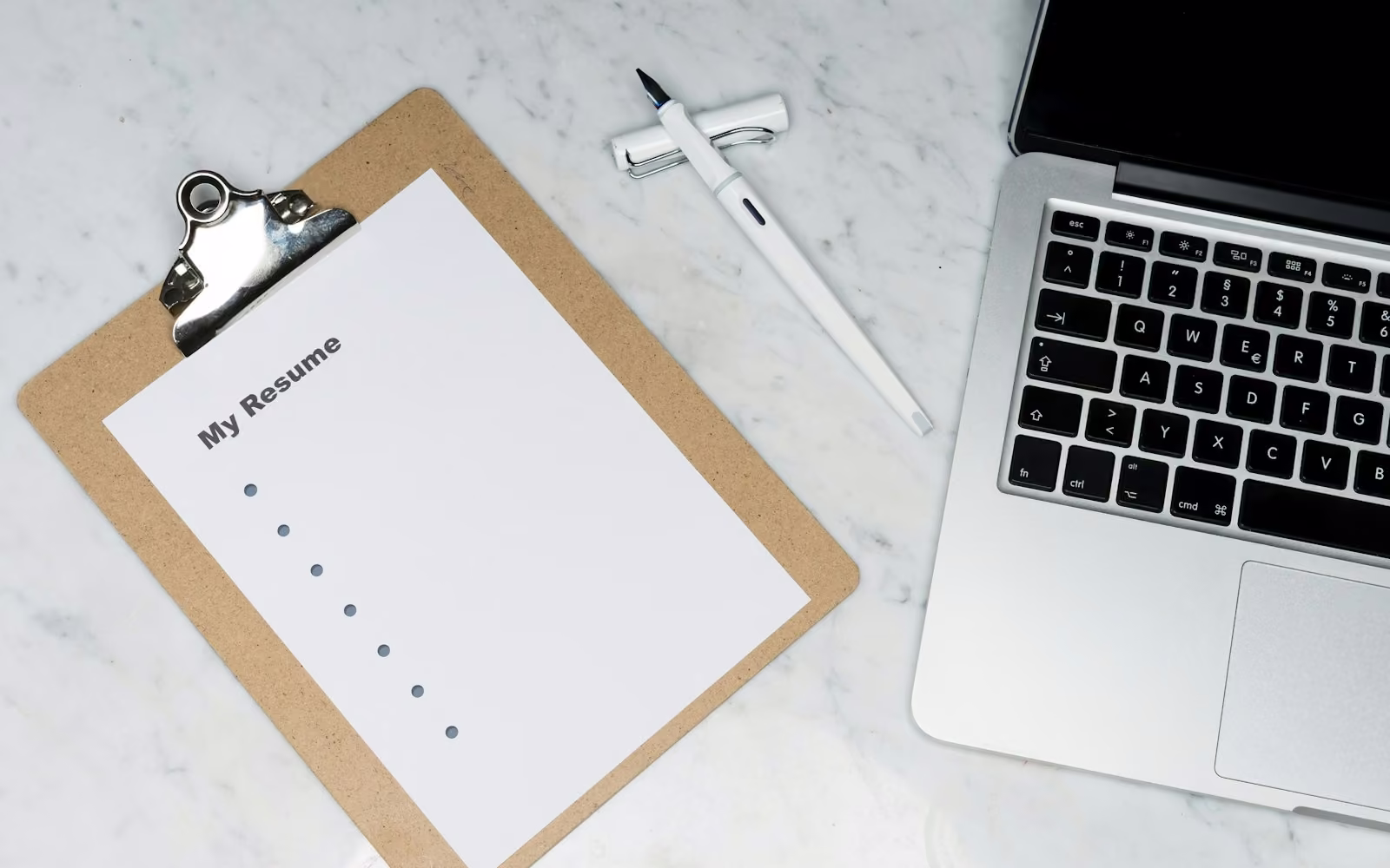 Papan jepit berisi kertas bertuliskan My Resume di samping laptop