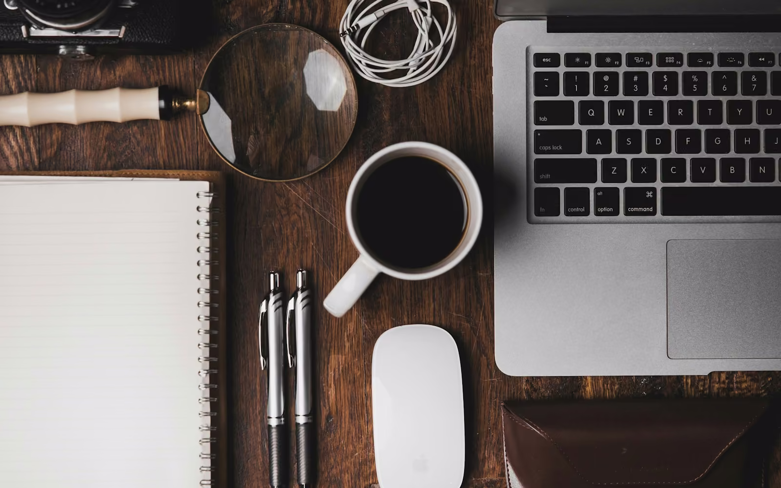 Meja kayu dengan laptop, buku catatan, kaca pembesar, dan secangkir kopi